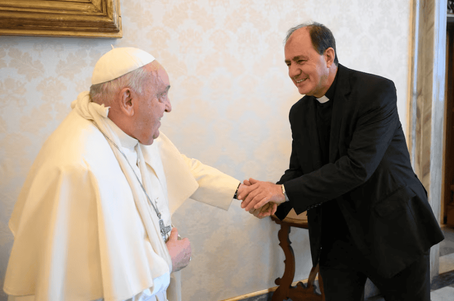 Papa Francisco saludando a un sacerdote en un encuentro formal, en un ambiente de cordialidad y respeto, con fondo de decoración sencilla y ambiente religioso.