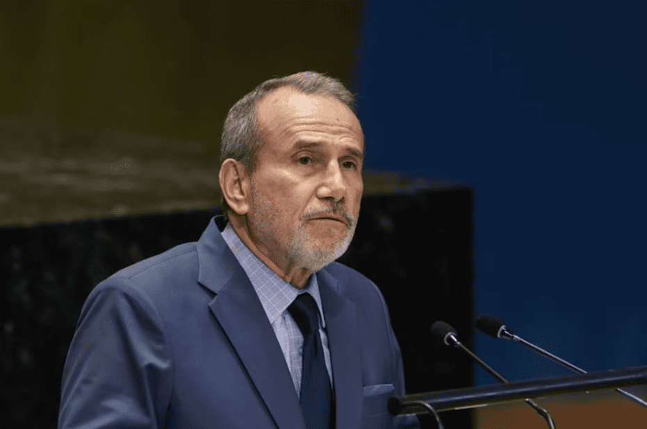 El hombre de edad avanzada con barba y cabello peinado hacia atrás, vestido con traje azul y corbata, hablando en un podio con micrófonos en un entorno formal.