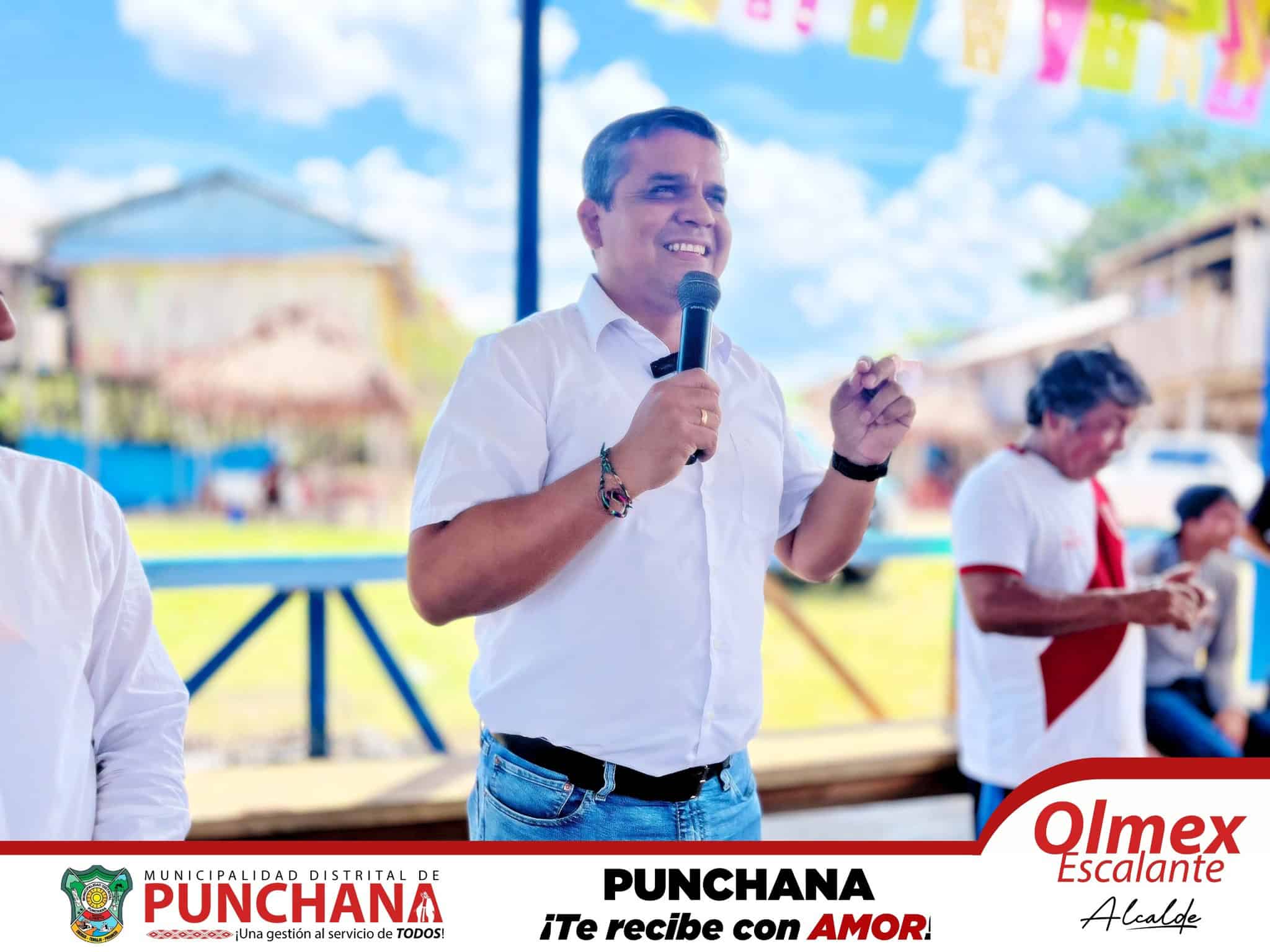 Aprobado hombre con micrófono en evento al aire libre, alcalde en la municipalidad de Punchana, participando en actividad comunitaria con un fondo de cielo azul y estructuras tradicionales.