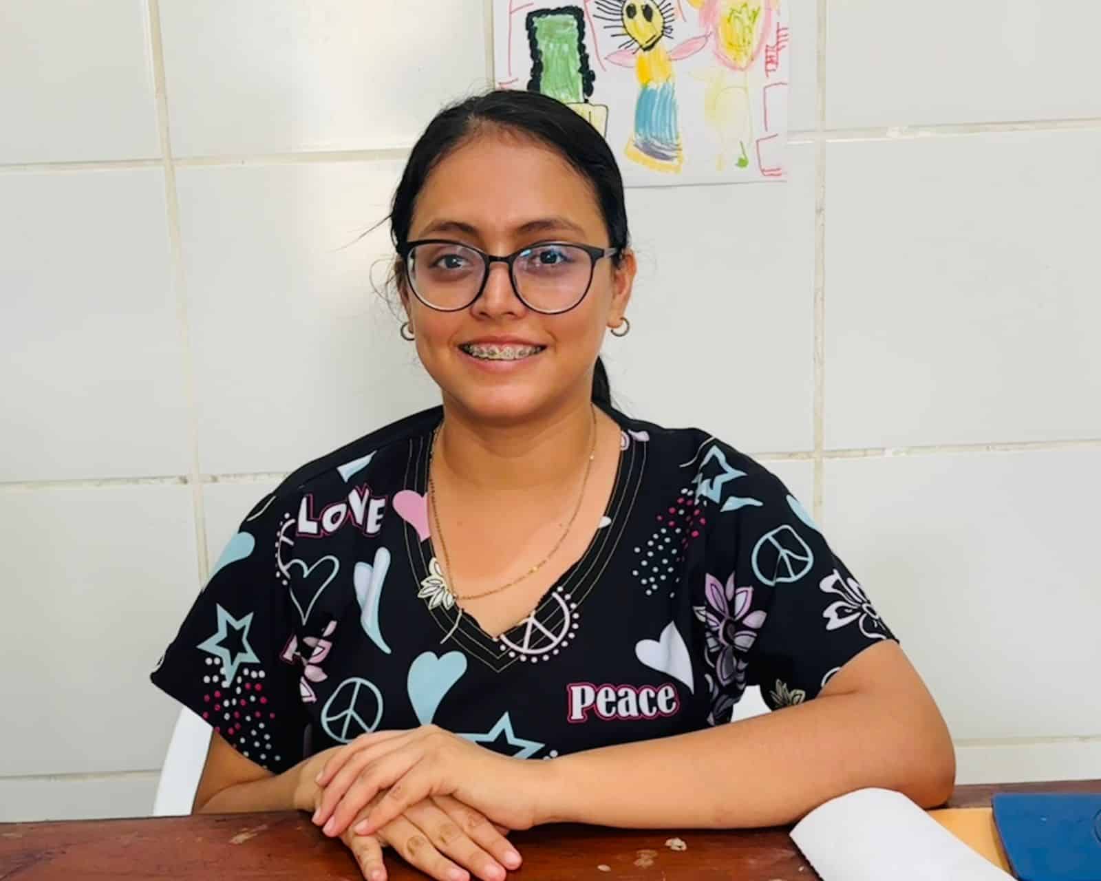 Niña con gafas y sonrisa, sentada en mesa en aula escolar, con dibujo infantil en la pared y libros en mesa, icono de paz y amor en su ropa, representando educación y alegría en Loreto Noticias.