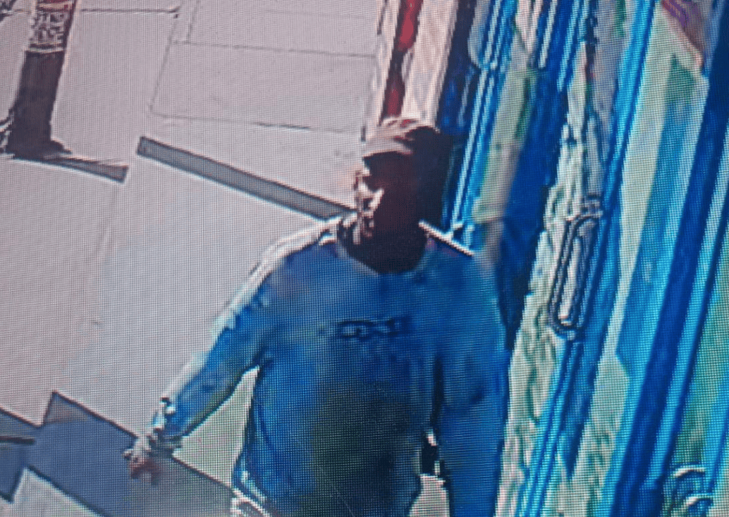 Hombre con chaqueta gris y camiseta azul oscuro en una tienda, sosteniendo una bolsa y usando una máscara facial, envuelto en un ambiente con paredes decoradas con patrones azules y beige.