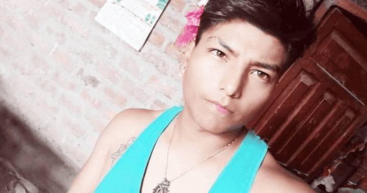 Joven con peinado estilo punk en tonos oscuros, usando camiseta sin mangas azul y collar de cadena con colgante, en ambiente casero con paredes de ladrillo y calendario en el fondo.