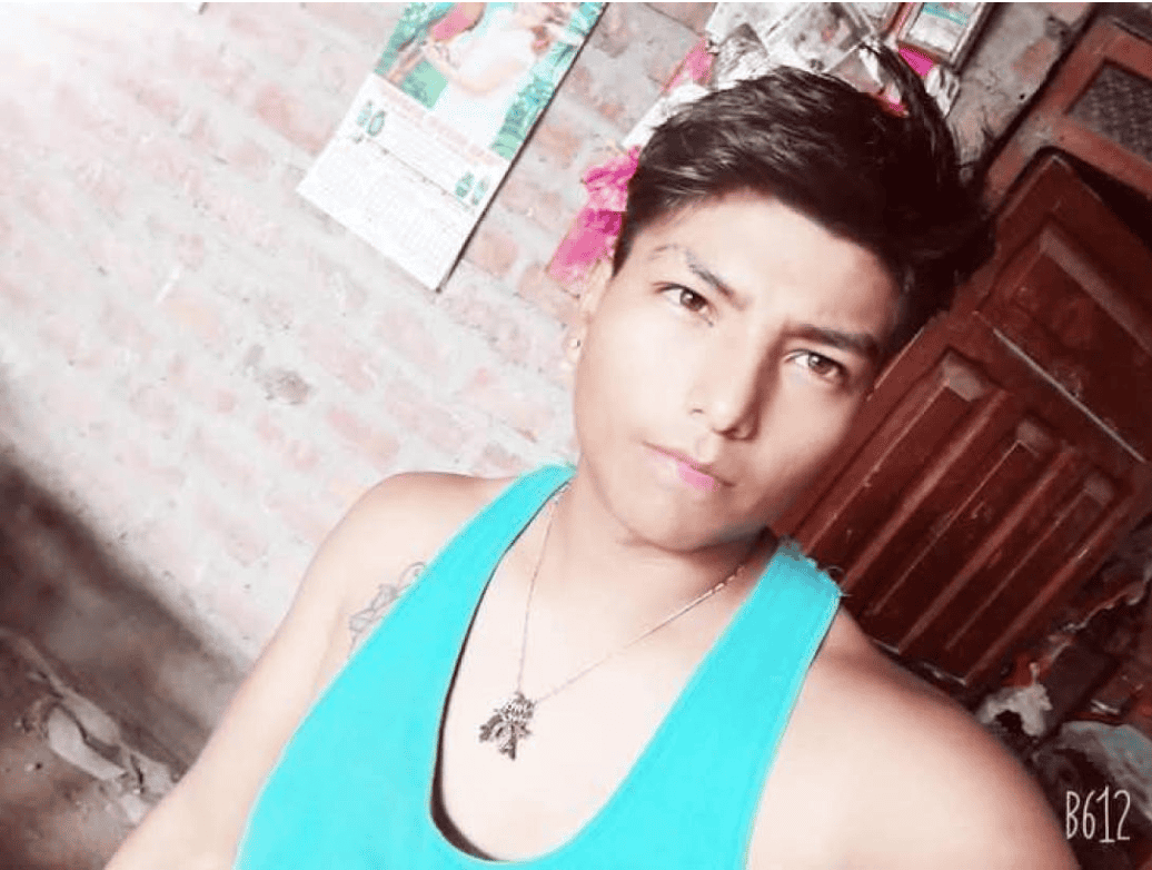 Joven con peinado estilo punk en tonos oscuros, usando camiseta sin mangas azul y collar de cadena con colgante, en ambiente casero con paredes de ladrillo y calendario en el fondo.