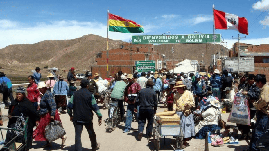 Bolivianos cruzando frontera hacia Perú en la entrada del paso fronterizo con banderas de Bolivia y Perú, en un día soleado con montañas de fondo y movimiento de personas y vehículos.