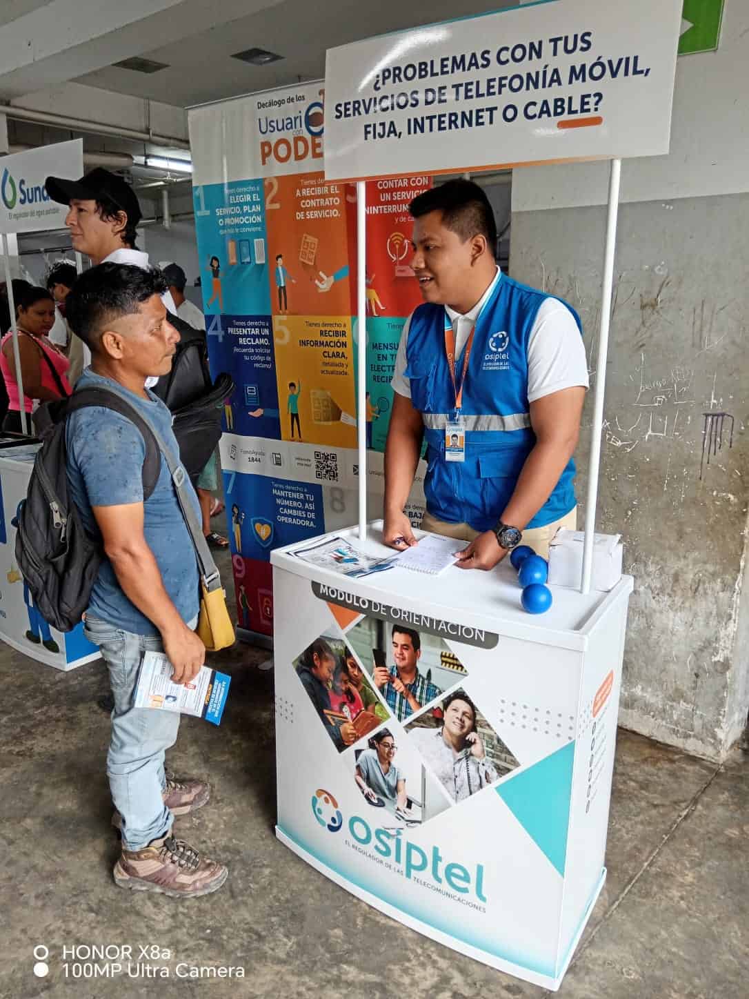 Accesorios de telecomunicaciones en feria de servicios móviles, internet y cable en Loreto, Perú. Usuarios reciben orientación sobre planes y contratos con OSIPTEL en evento informativo.