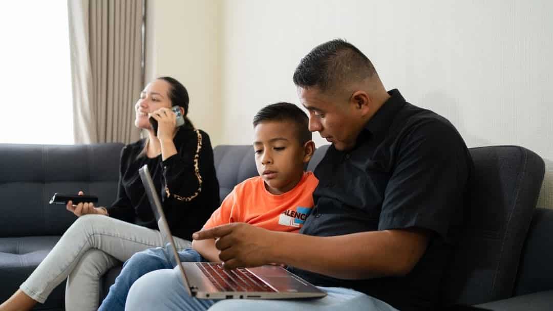 Padre y niño usando dispositivos electrónicos en el salón de su casa, mientras la madre escucha atención en teléfono móvil, ambiente familiar y moderno.