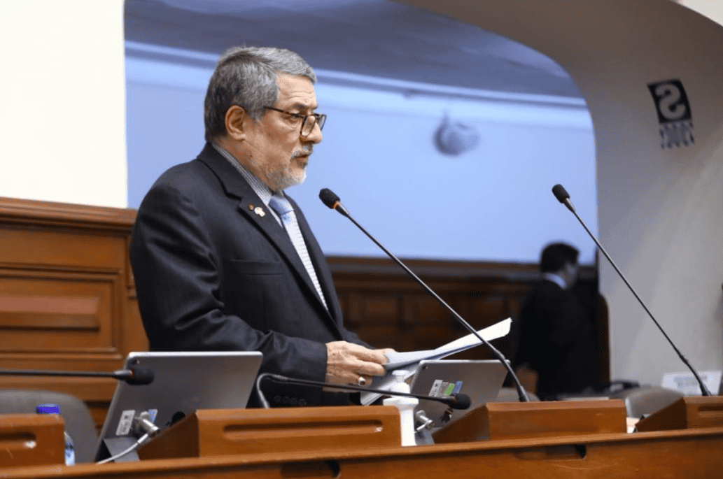 Consejo legislativo peruano en sesión plenaria, legislador en discurso con micrófono, ambiente formal, política y decisiones oficiales en Perú.