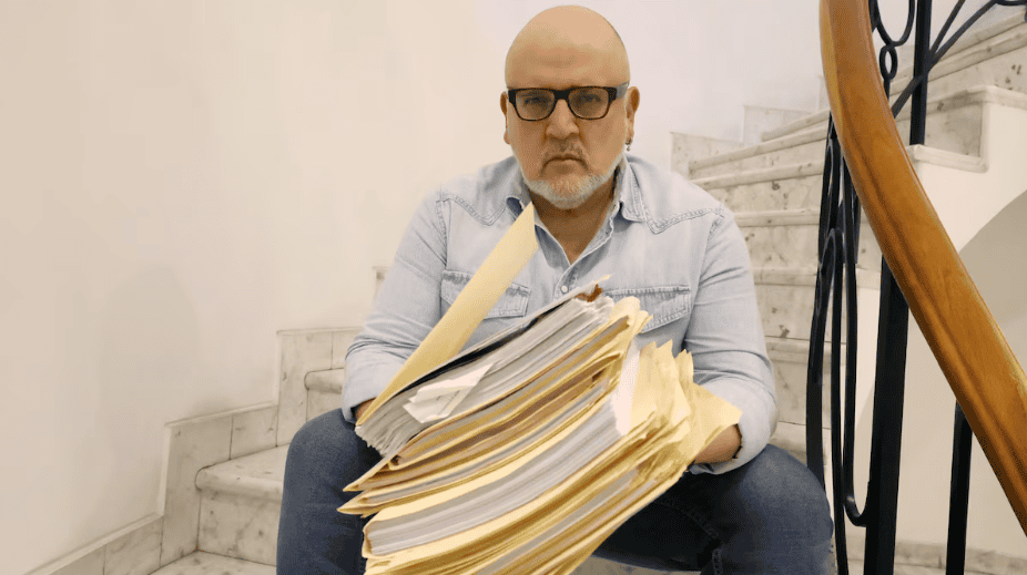 Archivador lleno de papeles y documentos, hombre con gafas y barba, expresión de frustración, escaleras de mármol, interior de edificio, situación de estrés laboral, noticias Loreto Noticias.
