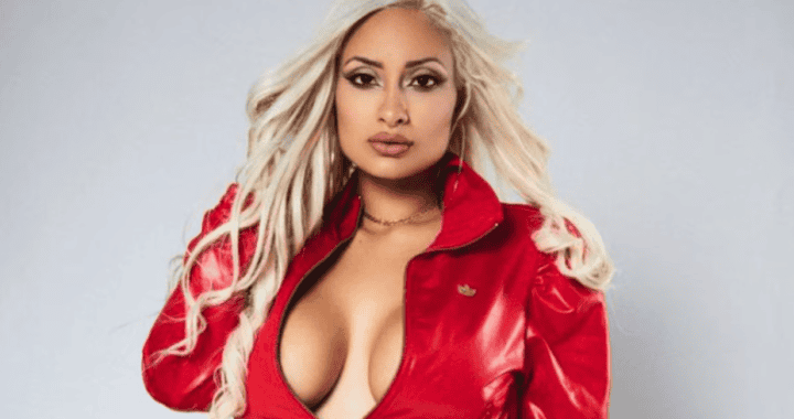 Chica con cabello rubio y peinado suelto, vestida con chaqueta roja y blusa ajustada, posando con expresión segura y mirada directa en fondo gris claro.