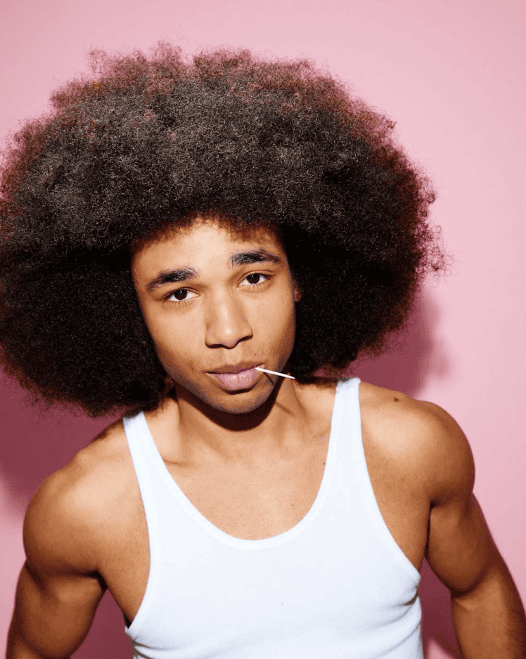 Amigo con cabello afro y camisa sin mangas posando en fondo rosa.