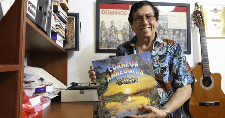 Mezcla de instrumentos musicales y libros, fotografía de un hombre sonriendo con una guitarra y un álbum en un ambiente de oficina con paredes decoradas con marcos y fotos, estilo cultural peruano.