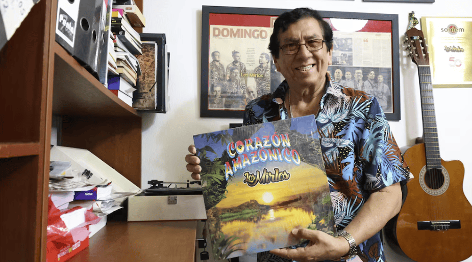 Mezcla de instrumentos musicales y libros, fotografía de un hombre sonriendo con una guitarra y un álbum en un ambiente de oficina con paredes decoradas con marcos y fotos, estilo cultural peruano.