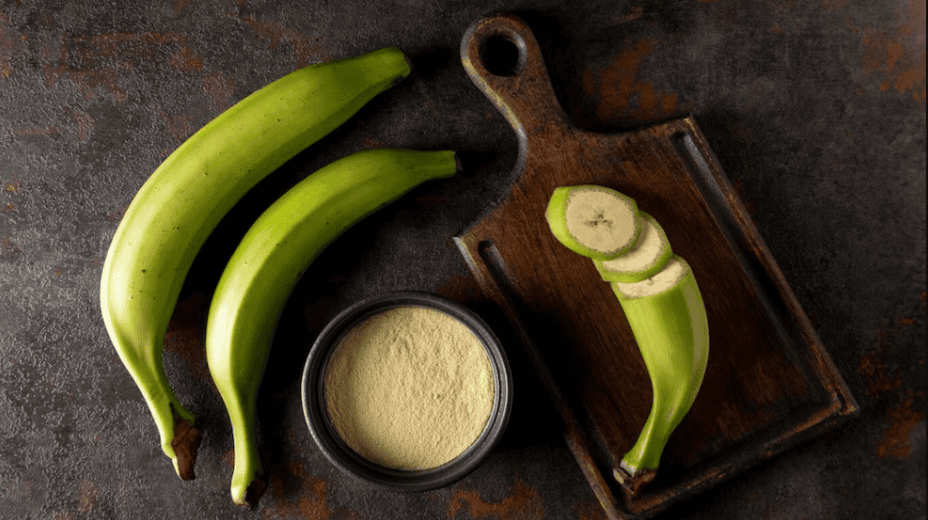 Bananos verdes y polvo de harina de plátano en una mesa de madera oscura.