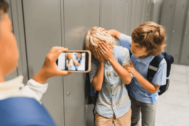 Joven ayudando a niño con queja en pasillo escolar, mientras otro toma una foto de la escena.