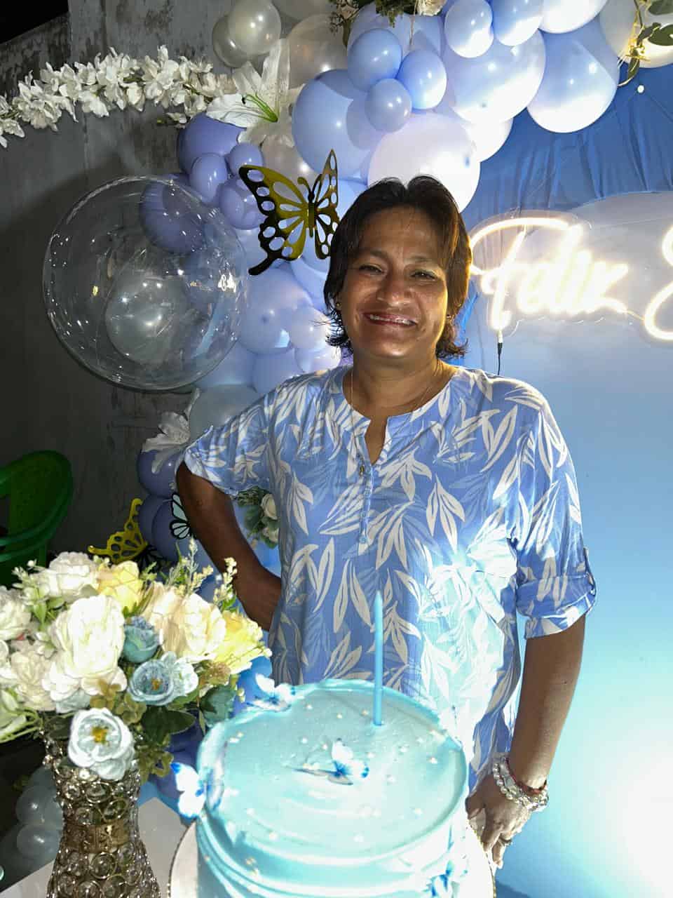 Fiesta de cumpleaños con decoración en tonos azules, globos y flores, celebrando a una mujer sonriente junto a un pastel temático con mariposas y un globo con la palabra "Felicidades".