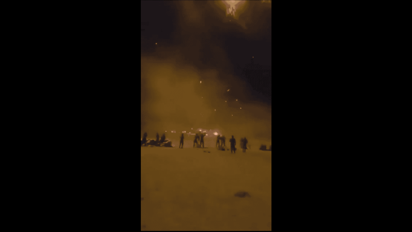 Balas de fuego y personas en la playa durante una noche con cielo oscuro y nubes.