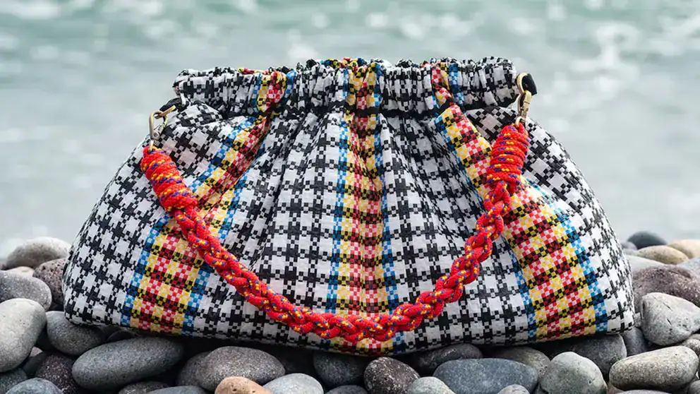 Alto texto: Bolsa artesanal de tejido con cuerda trenzada en diseño colorido, perfecta para accesorios de moda y cultura peruana, en un entorno de piedras junto al agua.