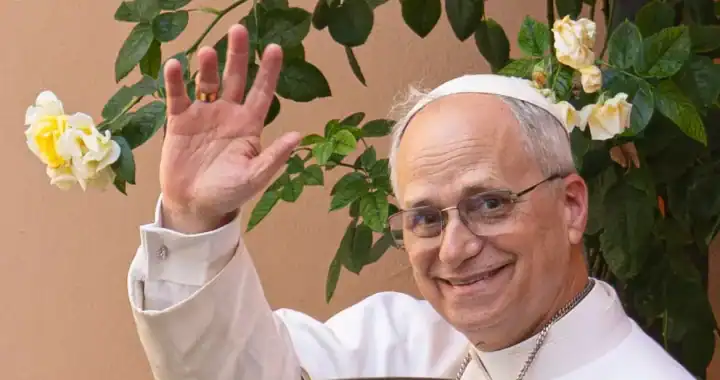 Líder religioso en una ceremonia, saludando y sonriendo, con flores blancas y verdes en el fondo.