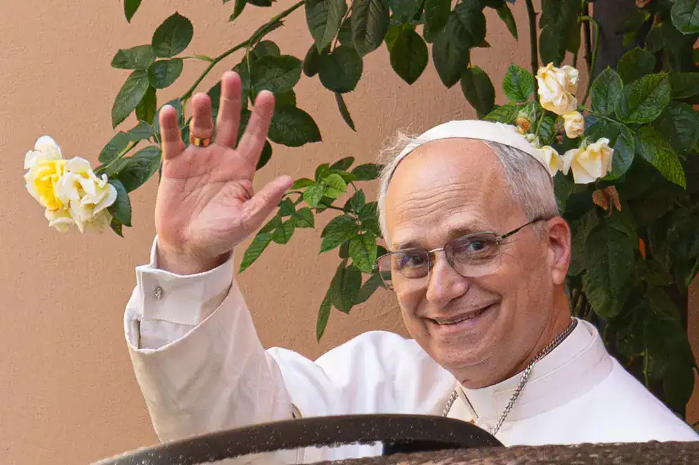 Líder religioso en una ceremonia, saludando y sonriendo, con flores blancas y verdes en el fondo.