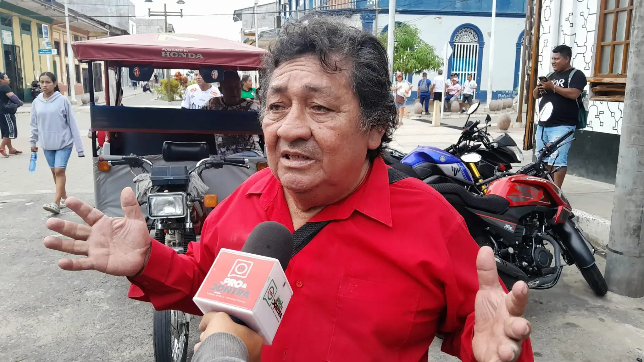 Camisa roja de un hombre siendo entrevistado en la calle de Loreto Noticias, en un ambiente urbano con motocicletas, personas caminando y edificio histórico al fondo.