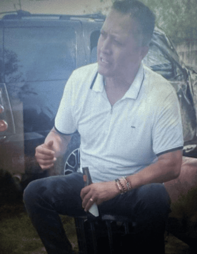 Persona hablando en un evento al aire libre, sentado en una silla, usando camiseta blanca con detalles oscuros y accesorios en la muñeca, con vehículos y naturaleza de fondo - cobertura de noticias locales Loreto Noticias en Perú.