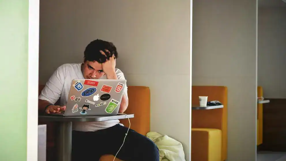 Estudiante frustrado usando laptop con stickers de programación y desarrollo en un espacio de estudio.