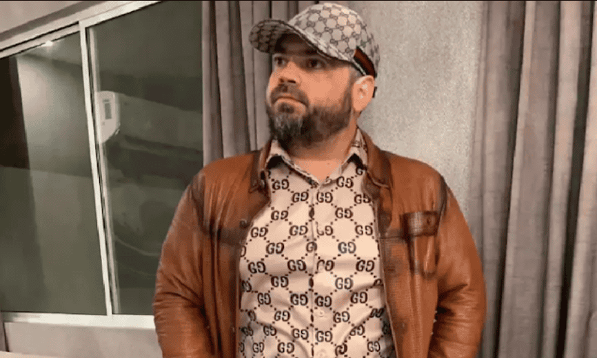 Guerrero con gafas y barba viste chaqueta de cuero y camisa con logo de Gucci, posando en un espacio interior con paredes de tela gris.