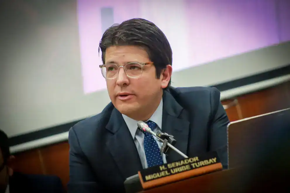Hombre en una reunión formal hablando en un evento oficial, usando traje y gafas, con un micrófono frente a él y nombre en placa Diplomático.