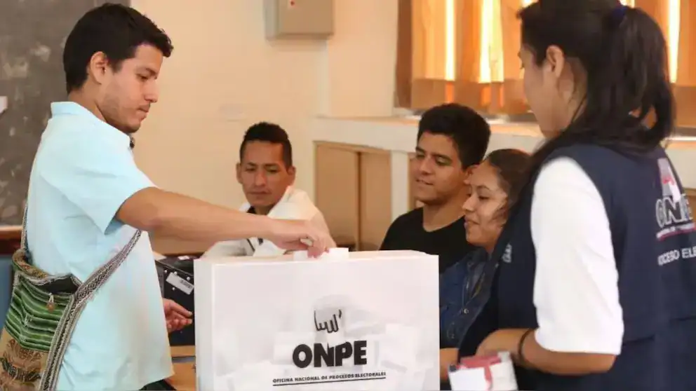 Votación en Loreto durante proceso electoral en Perú, persona depositando su voto en urna con miembros del comité electoral y periodistas presentes.