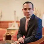 Hombre en traje formal con banderas de Perú en fondo, en una oficina gubernamental.