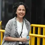 Una mujer sonriente con cabello rizado, vestida con blusa blanca y suéter gris, sostiene un agua y está en un entorno urbano con barreras amarillas, en Loreto Noticias.