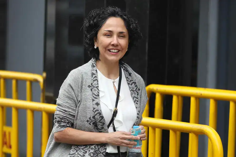 Una mujer sonriente con cabello rizado, vestida con blusa blanca y suéter gris, sostiene un agua y está en un entorno urbano con barreras amarillas, en Loreto Noticias.