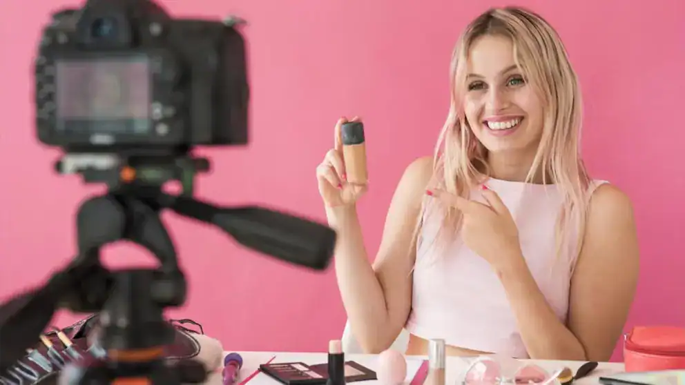 Mujer sonriendo mostrando producto cosmético con fondo rosa, en un video de belleza y maquillaje, en un entorno colorido y alegre para tutoriales y reseñas de cosméticos.