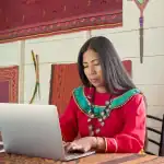 Mujer indígena trabajando en computador en comunidad peruana con artefactos y tejidos tradicionales.