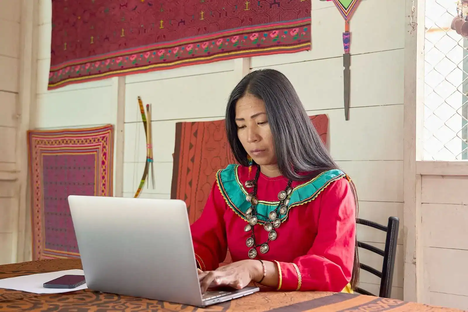 Mujer indígena trabajando en computador en comunidad peruana con artefactos y tejidos tradicionales.